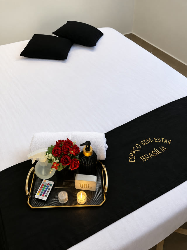 Maca com lençol branco, faixa preta bordada em dourado com o nome do espaço e bandeja com rosas, vela e óleo
