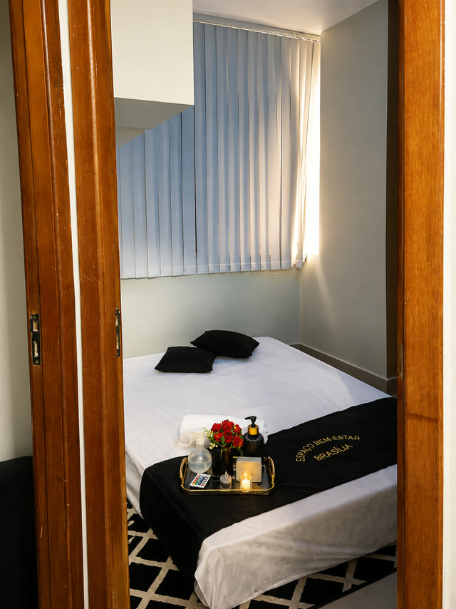 Sala de atendimento vista da porta, com maca arrumada, almofadas pretas e luz natural pela persiana